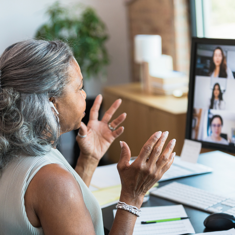 A woman with gray hair participates in a video call, gesturing with her hands while four people appear on her computer screen in a home office setting.