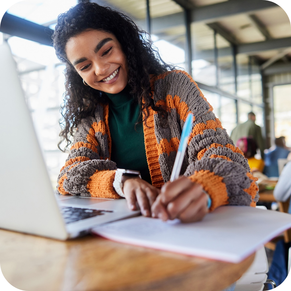 A woman sits at a table, smiling while writing in a notebook and using a laptop in a bright, modern indoor space.
