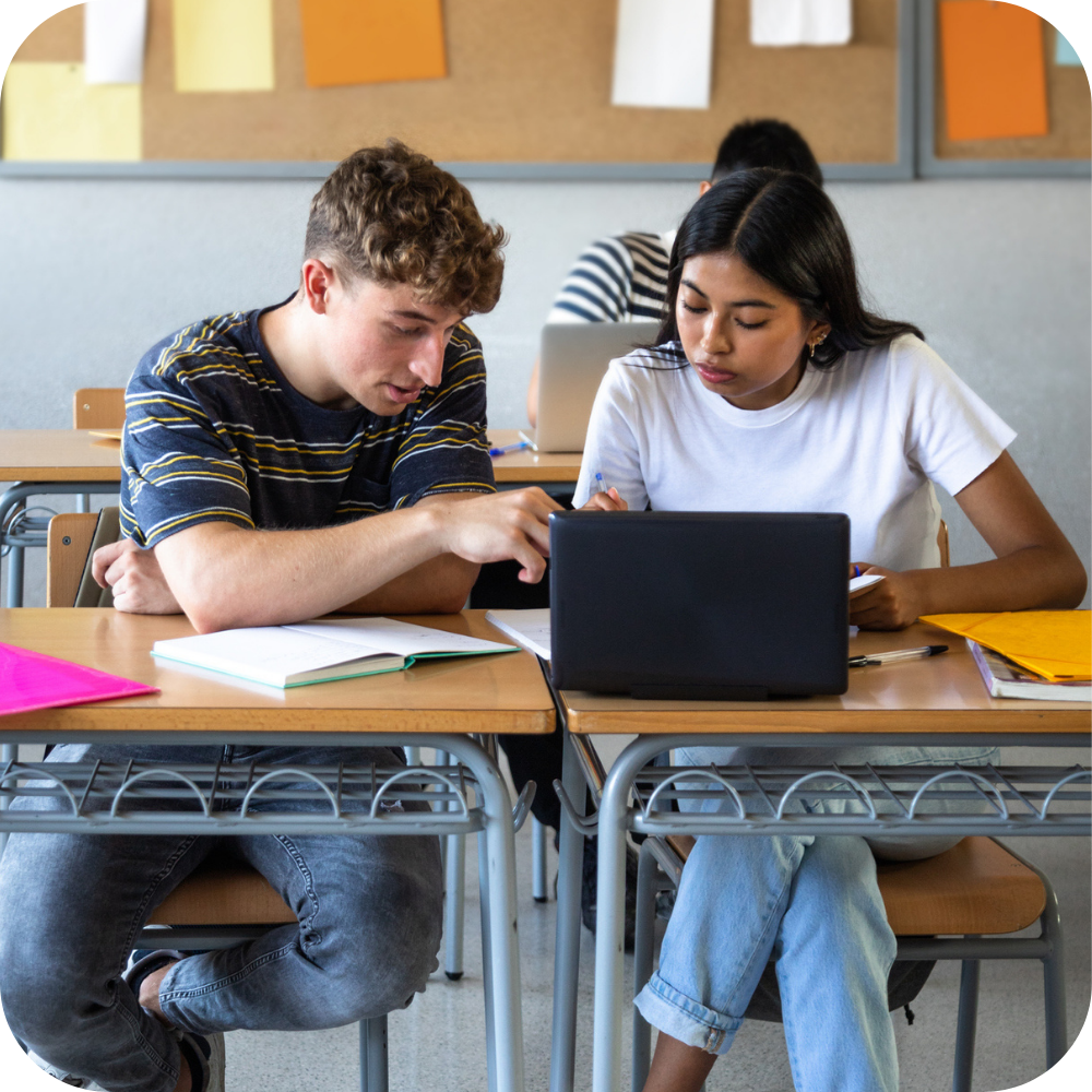 Two students sit at desks in a classroom, working together on a laptop with notebooks and papers spread out in front of them.