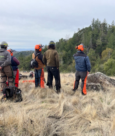 A group of people wearing protective gear, including helmets and ear protection, stand in a grassy field facing a forested hillside.