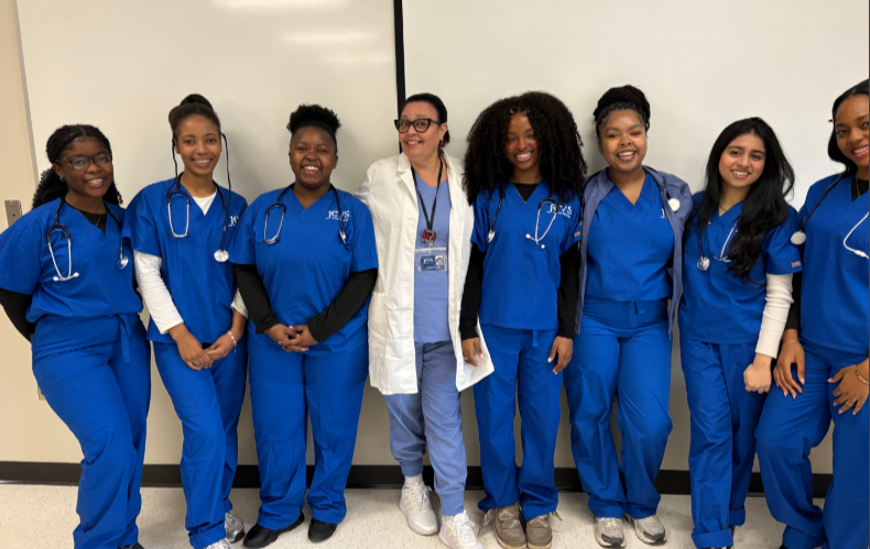 Eight women, seven in blue scrubs and one in a white lab coat, stand together and smile in front of a whiteboard in a clinical setting.
