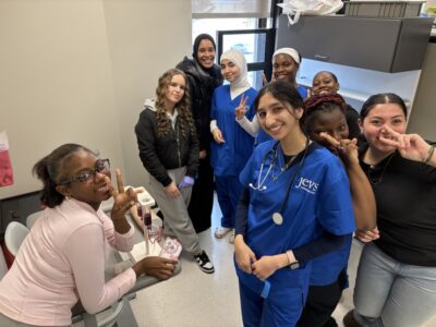 A group of students, some in blue medical scrubs and some in casual clothes, smile and pose together in a classroom or clinic setting.
