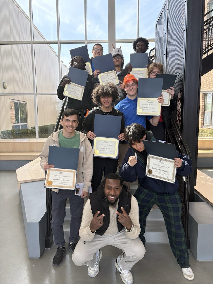 A group of students pose on a staircase, each holding a certificate. An adult kneels in front, making a peace sign. The setting appears to be indoors with large windows.