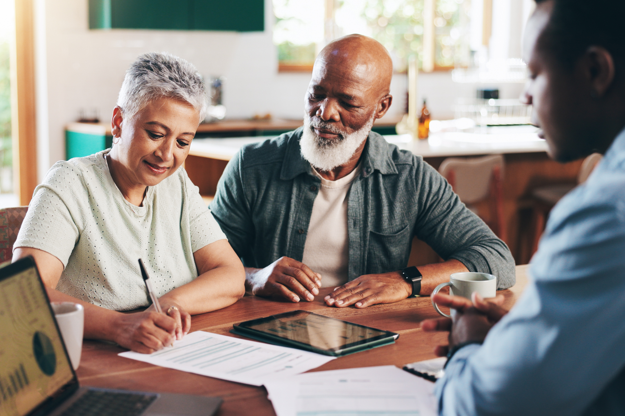 Three adults sit at a table with documents and a tablet; one woman signs paperwork while two men observe. A laptop and coffee mugs are also on the table.