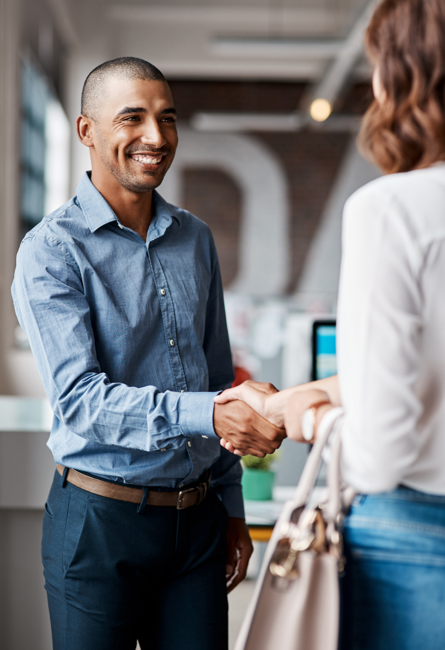 A man in a blue shirt smiles while shaking hands with a woman in an office setting.