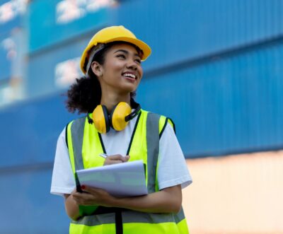 A woman wearing a hard hat, safety vest, and headphones stands outdoors, holding a notepad and pen, with shipping containers in the background.