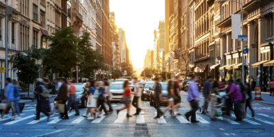 People crossing a busy city street at a crosswalk during sunset, with blurred motion indicating movement; tall buildings line both sides of the street.