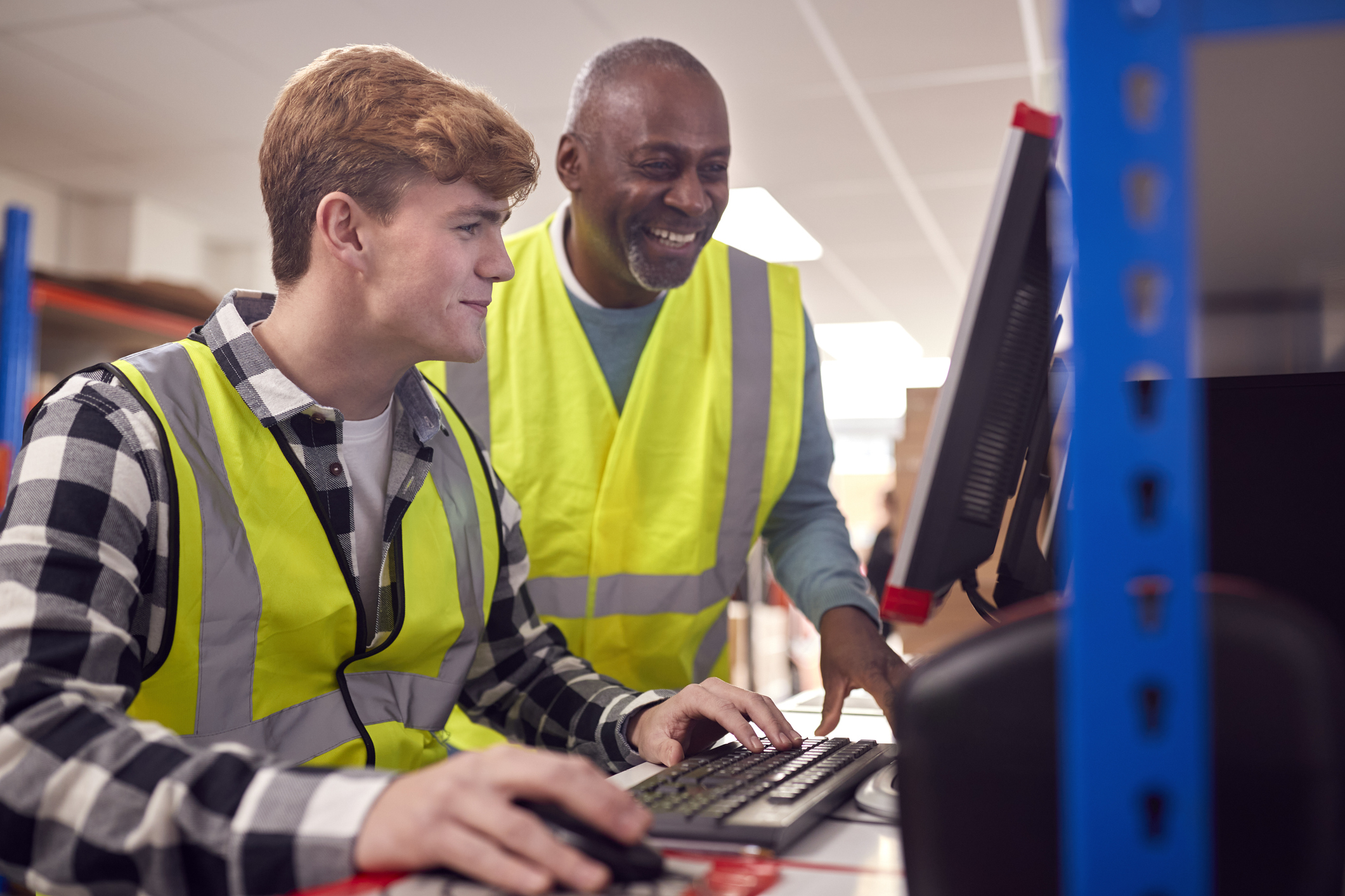 Two men wearing yellow safety vests work at a computer in a warehouse setting, with shelves and boxes visible in the background.