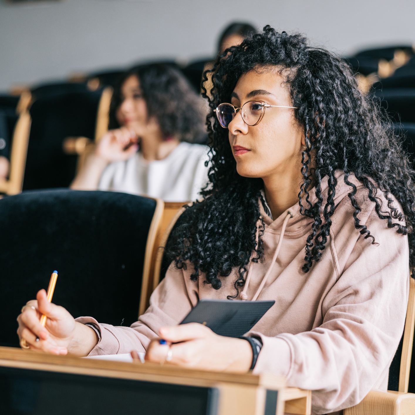A woman with curly hair and glasses sits in a lecture hall, holding a pen and tablet, attentively listening and taking notes. Other students are seated in the background.
