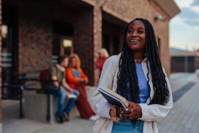 A young woman holding notebooks smiles while standing outside a brick building; three people sit and talk in the background.
