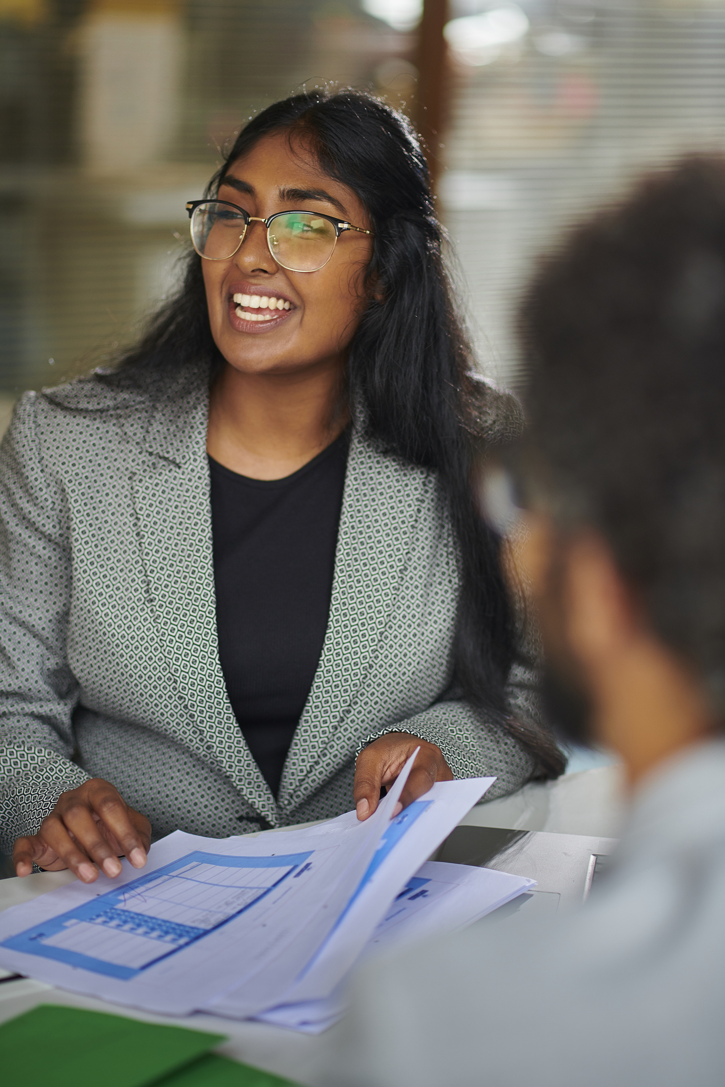 A woman in a gray blazer holds documents and smiles while talking to a person seated across from her in an office setting.