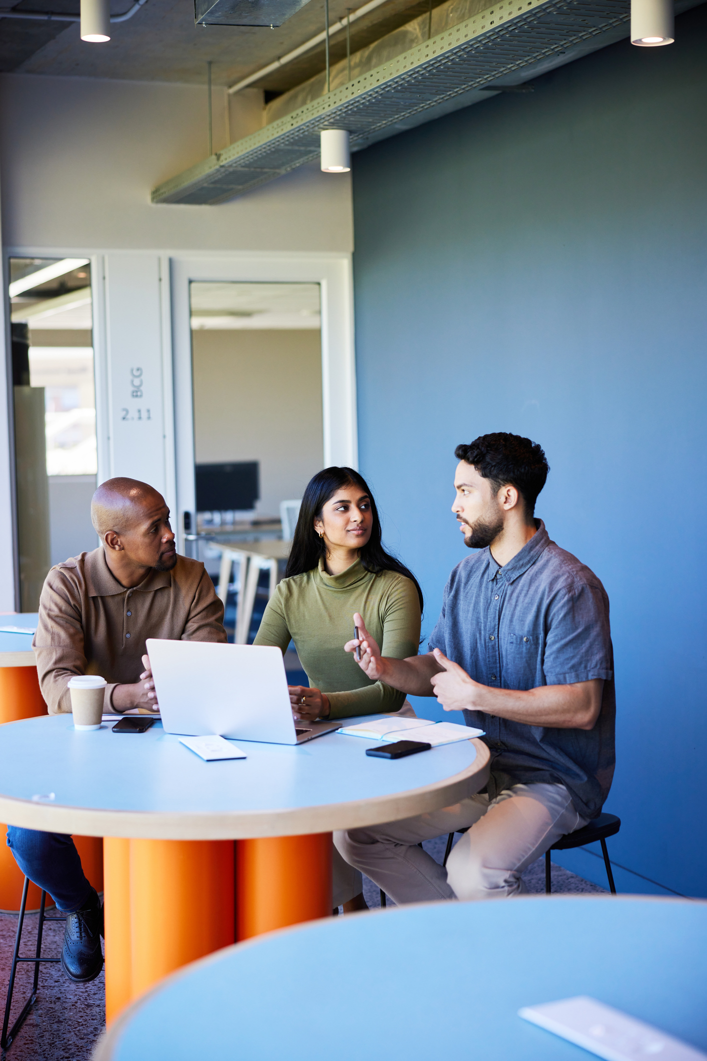 Three people sit at round tables with a laptop and smartphones, having a discussion in a modern office space with blue and orange decor.