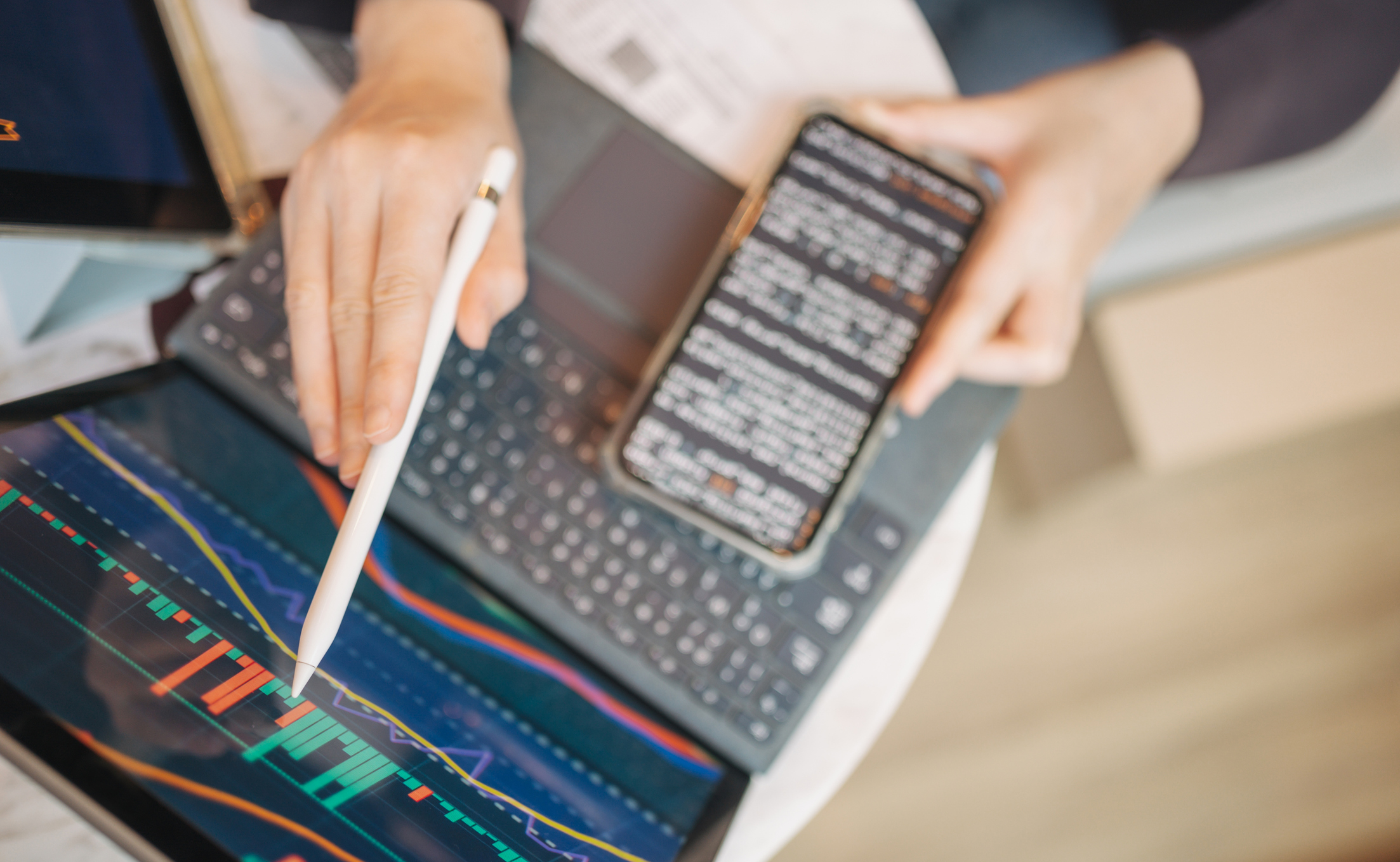 A person uses a stylus on a tablet displaying financial charts, while holding a smartphone with text on the screen and a keyboard nearby.