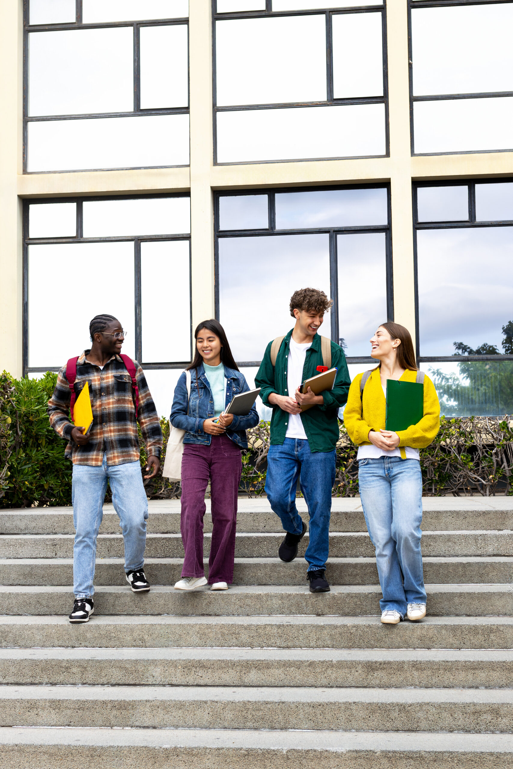 Four students with backpacks and books walk and talk together down outdoor steps in front of a modern building.