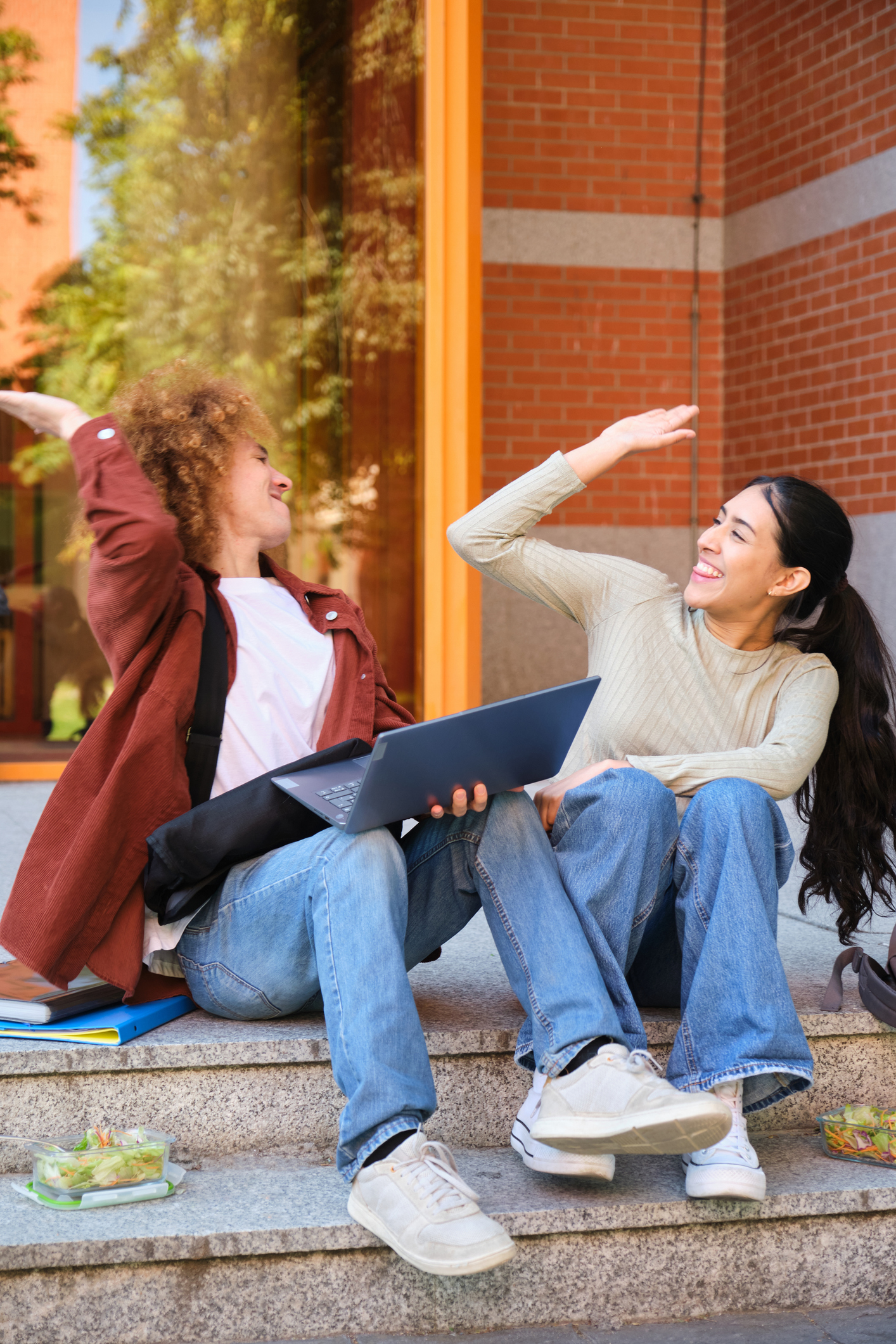 Two young adults sitting on outdoor steps with a laptop and salads, smiling and giving each other a high five.