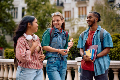Three students stand outdoors, smiling and talking. One holds books, another has headphones around their neck, and they all carry backpacks. Trees and buildings are visible in the background.