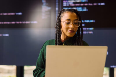 A person with braided hair and glasses works on a laptop, with computer code displayed on large screens in the background.