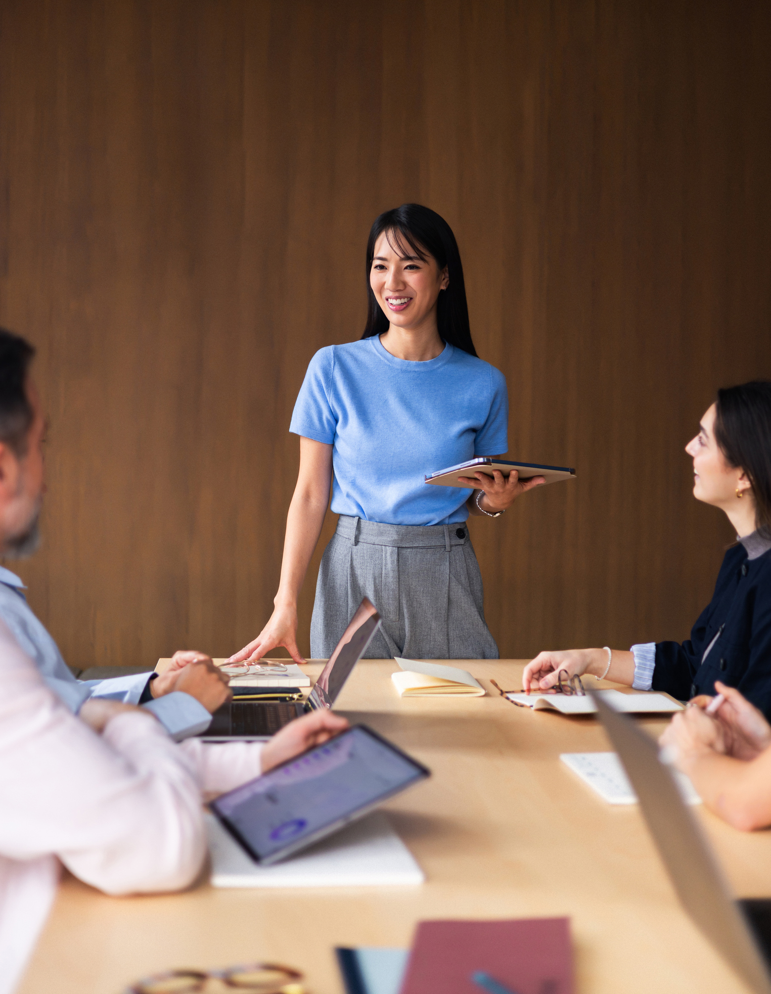 A woman stands at a table holding a tablet, speaking to three seated colleagues during a meeting in a modern office setting.