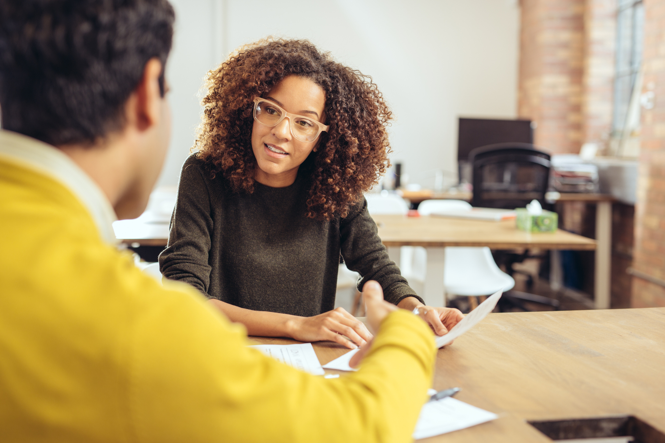 Two people sit at a desk in an office, engaged in a conversation. One person takes notes while listening attentively to the other.