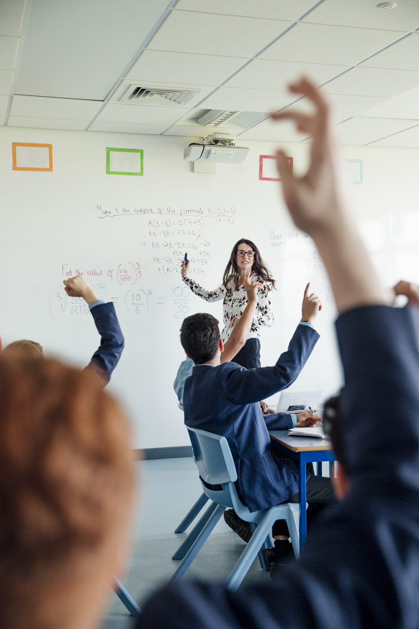 A teacher stands at a whiteboard explaining math equations while several students seated at desks raise their hands.