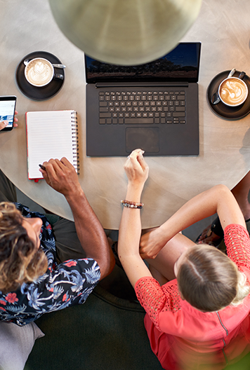Two people sit at a round table with a laptop, notebook, smartphone, and two cups of coffee, seen from above. One writes in the notebook while the other gestures toward the laptop.