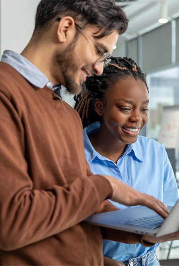 Two people stand together in an office, both smiling and looking at a laptop one of them is holding.
