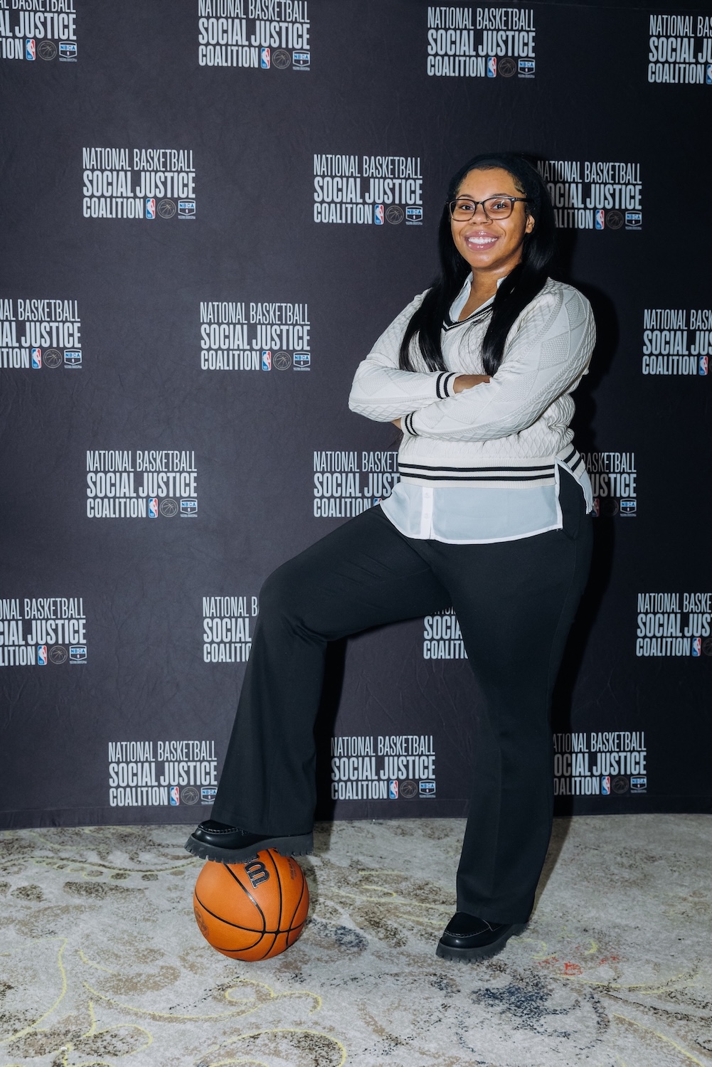 A woman in business attire stands with one foot on a basketball, posing in front of a