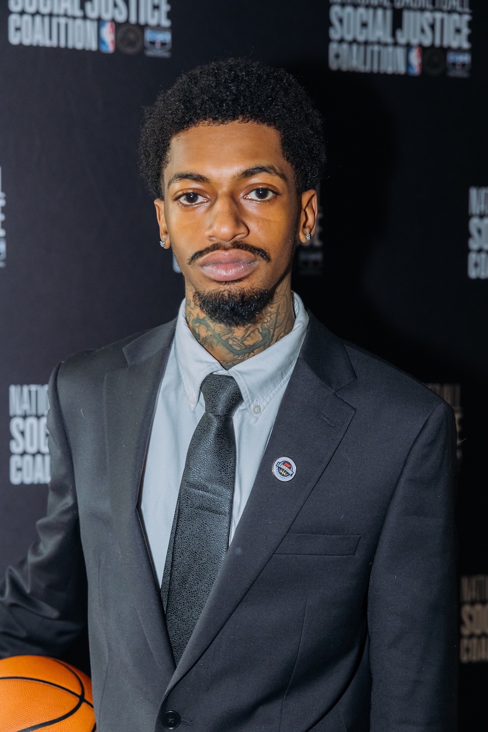 A man in a suit and tie holding a basketball stands in front of a backdrop with the words