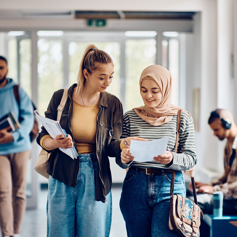 Two young women, one wearing a hijab, walk indoors holding papers and talking. Other people with books and laptops are visible in the background.