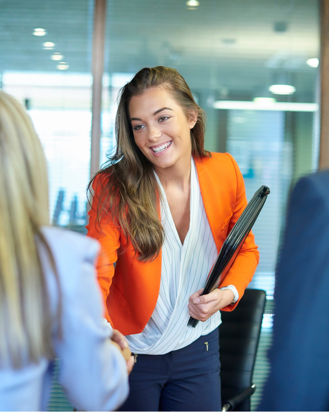 Woman in an orange blazer smiles and shakes hands with another person in an office setting, holding a folder under her arm.