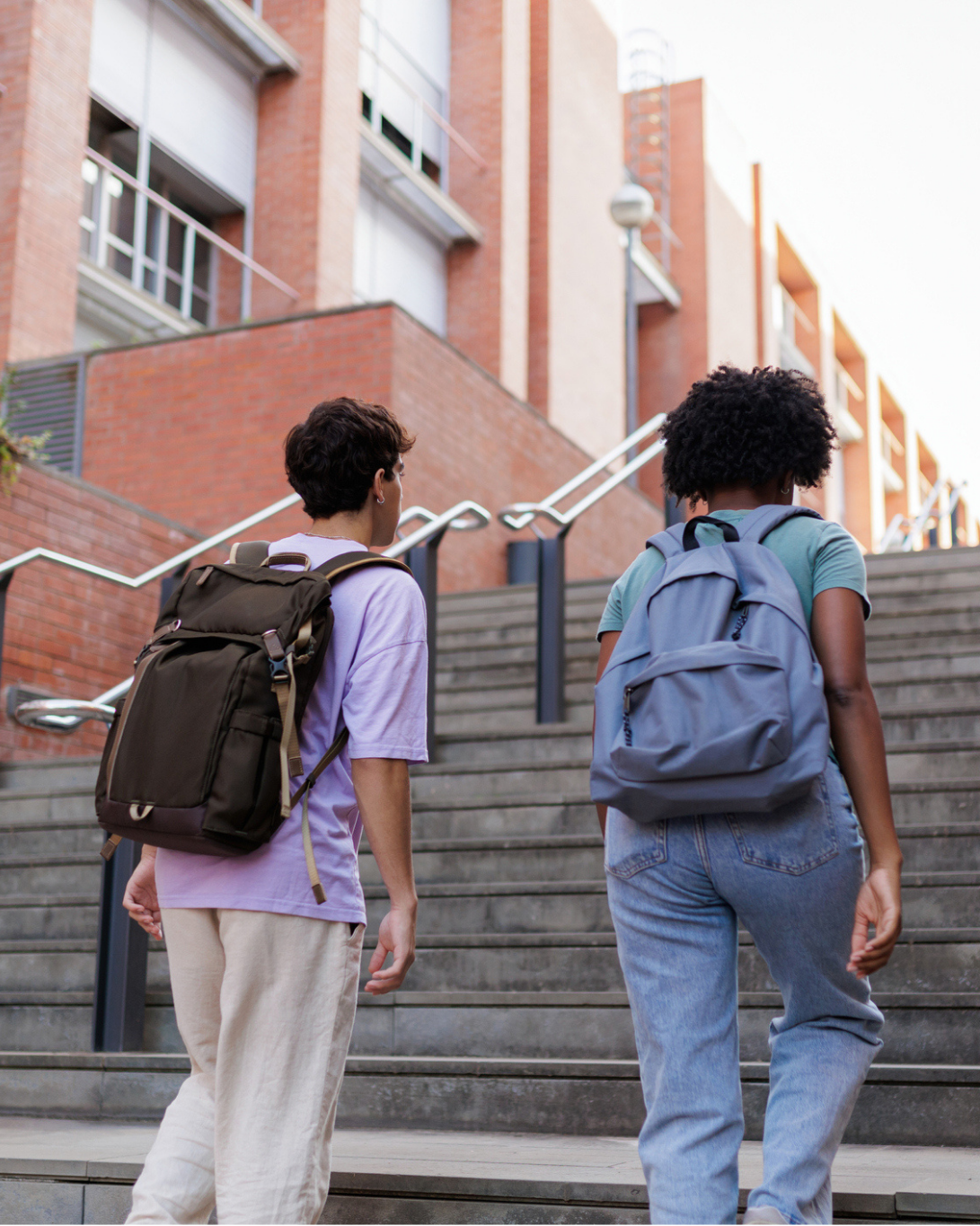 Two students with backpacks walk up outdoor stairs toward a modern brick school building.