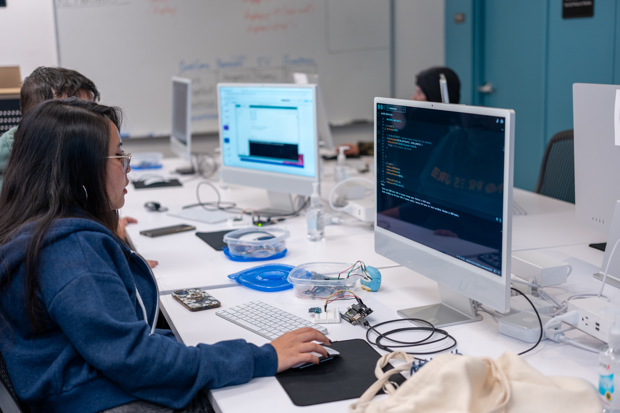 A person is seated at a desk using a computer with code on the screen in a classroom setting, alongside others working on similar tasks.