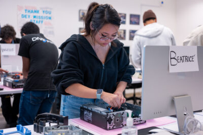 A woman assembles or repairs a computer in a classroom setting with other people working on similar tasks in the background.