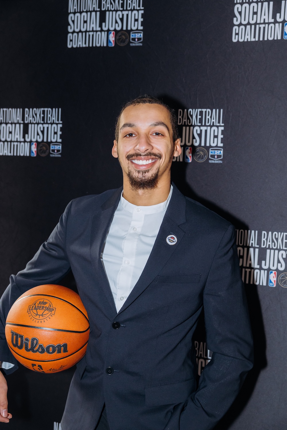 A man in a suit smiles and holds a Wilson basketball in front of a National Basketball Social Justice Coalition backdrop.
