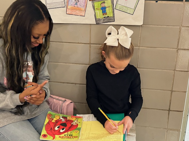 Two girls sit next to each other; one writes in a yellow notebook while the other observes. A book titled 