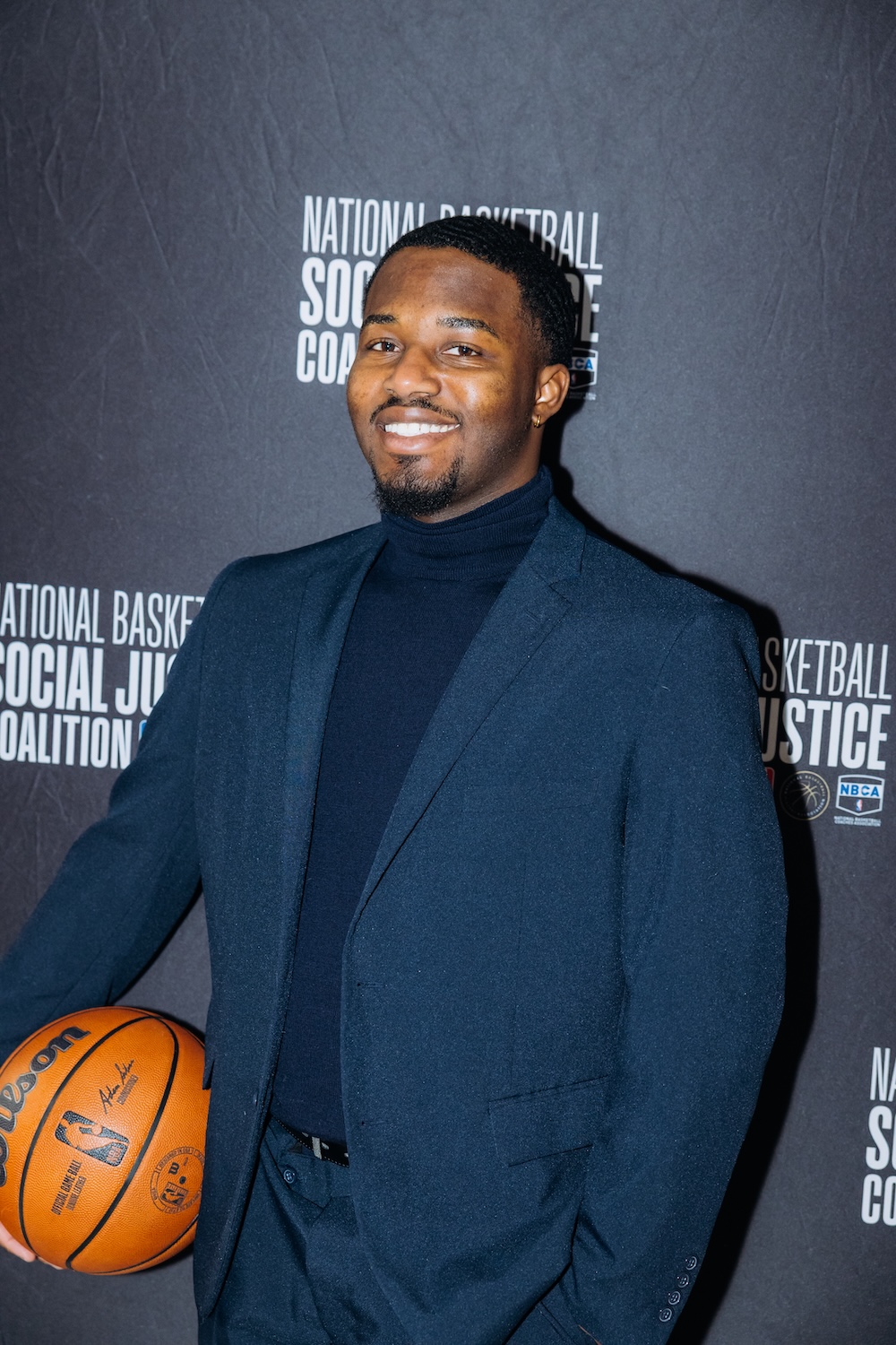 A man in a dark suit and black turtleneck stands holding a basketball in front of a backdrop that reads