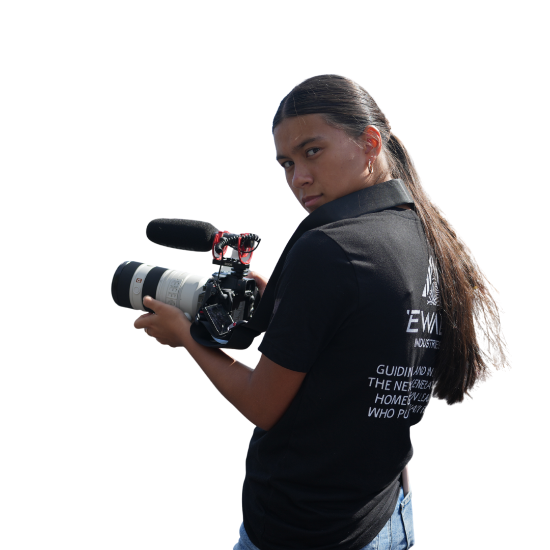 A person with long hair holds a professional camera with a microphone, wearing a black shirt with text on the back, facing toward the viewer.