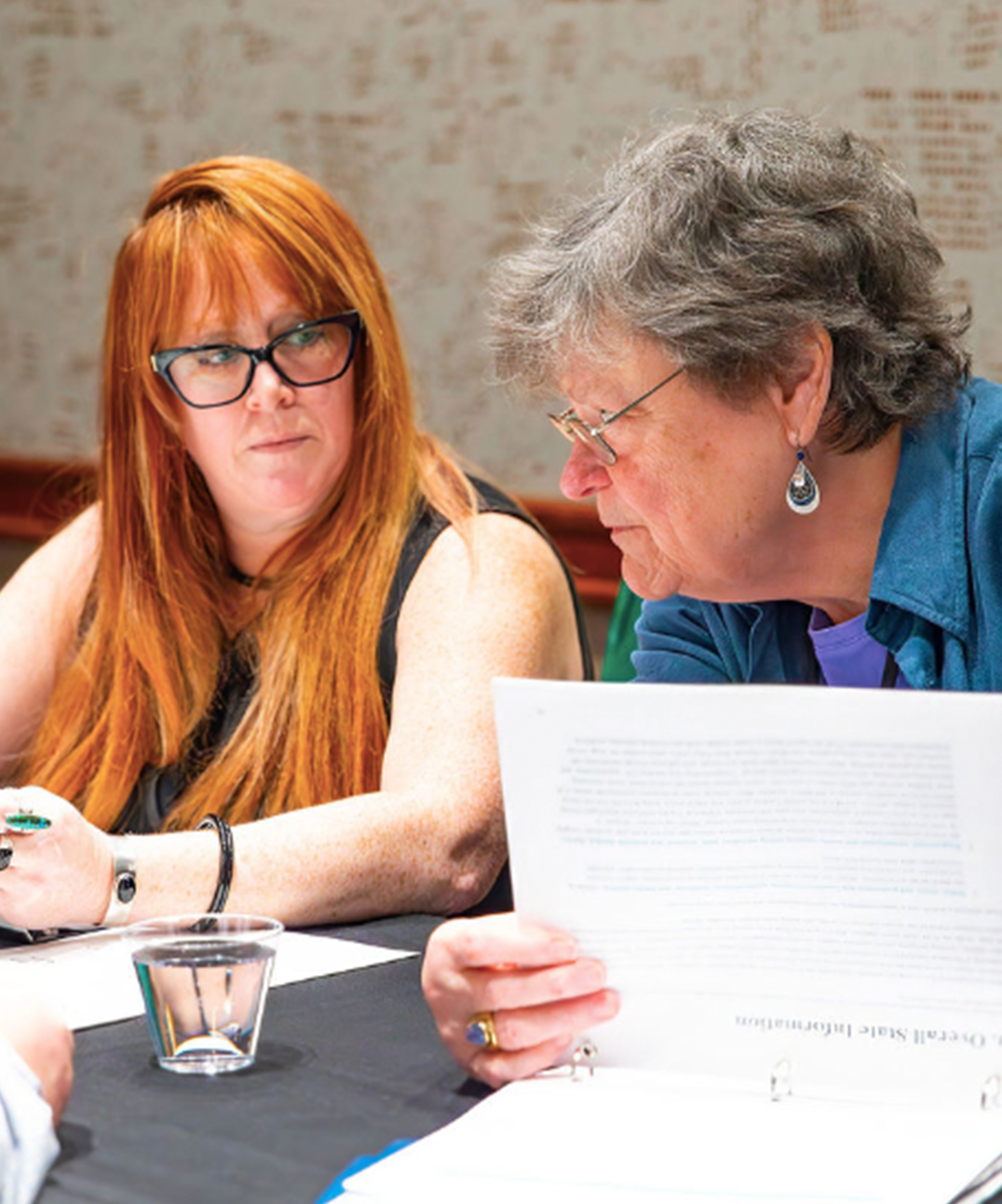 Two women sit at a table, one reading from a document while the other listens attentively. There are papers, a pen, and a glass of water on the table.