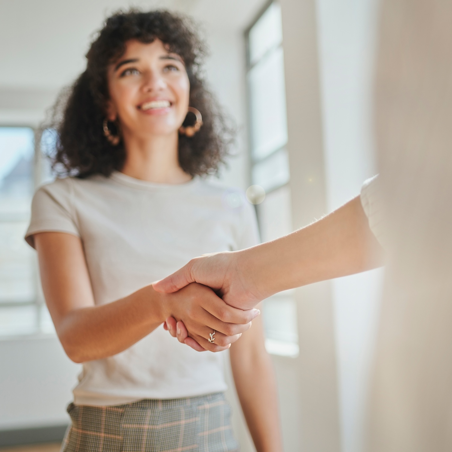 Smiling woman with curly hair and hoop earrings shakes hands with another person indoors, standing near a window.