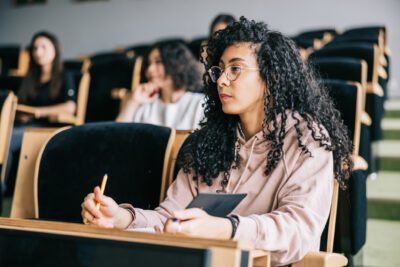 A student with curly hair and glasses sits in a lecture hall, holding a notebook and pencil, attentively listening. Several other students are seated in the background.