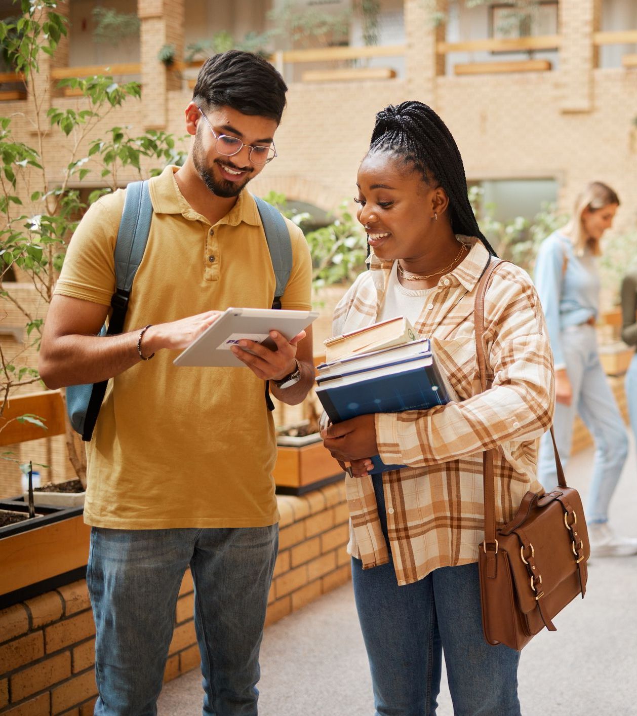 Two students stand indoors; one holds books while the other shows something on a tablet. Both are smiling and appear to be having a friendly conversation.