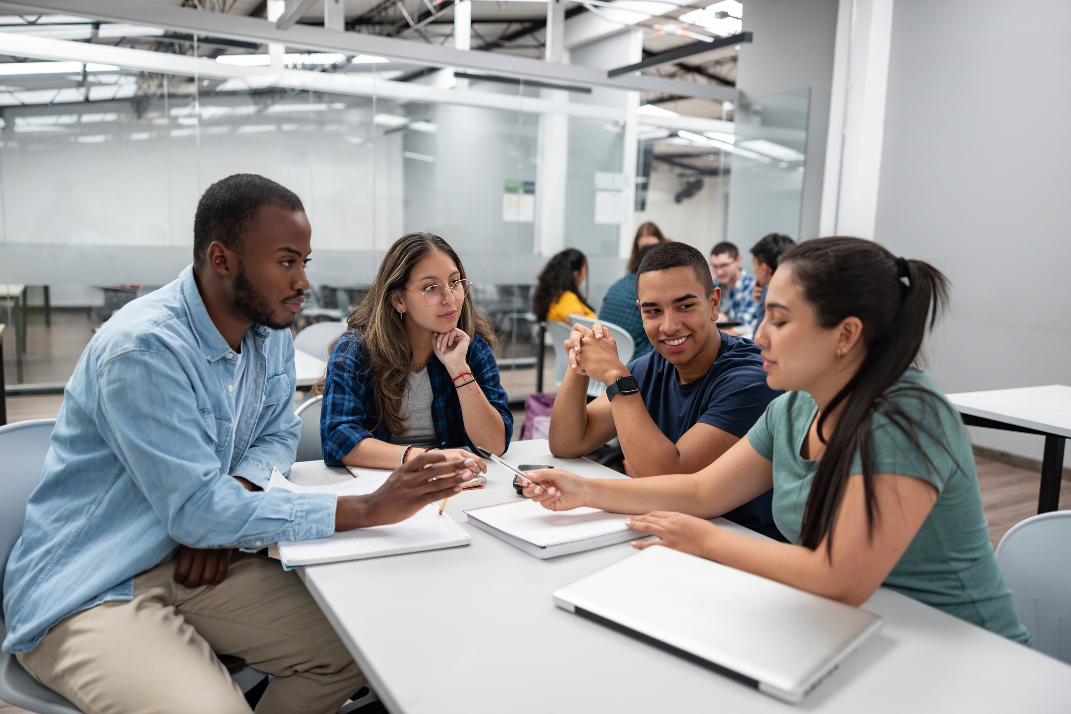 Four students sit at a table in a classroom, discussing and sharing notes with notebooks and laptops in front of them.