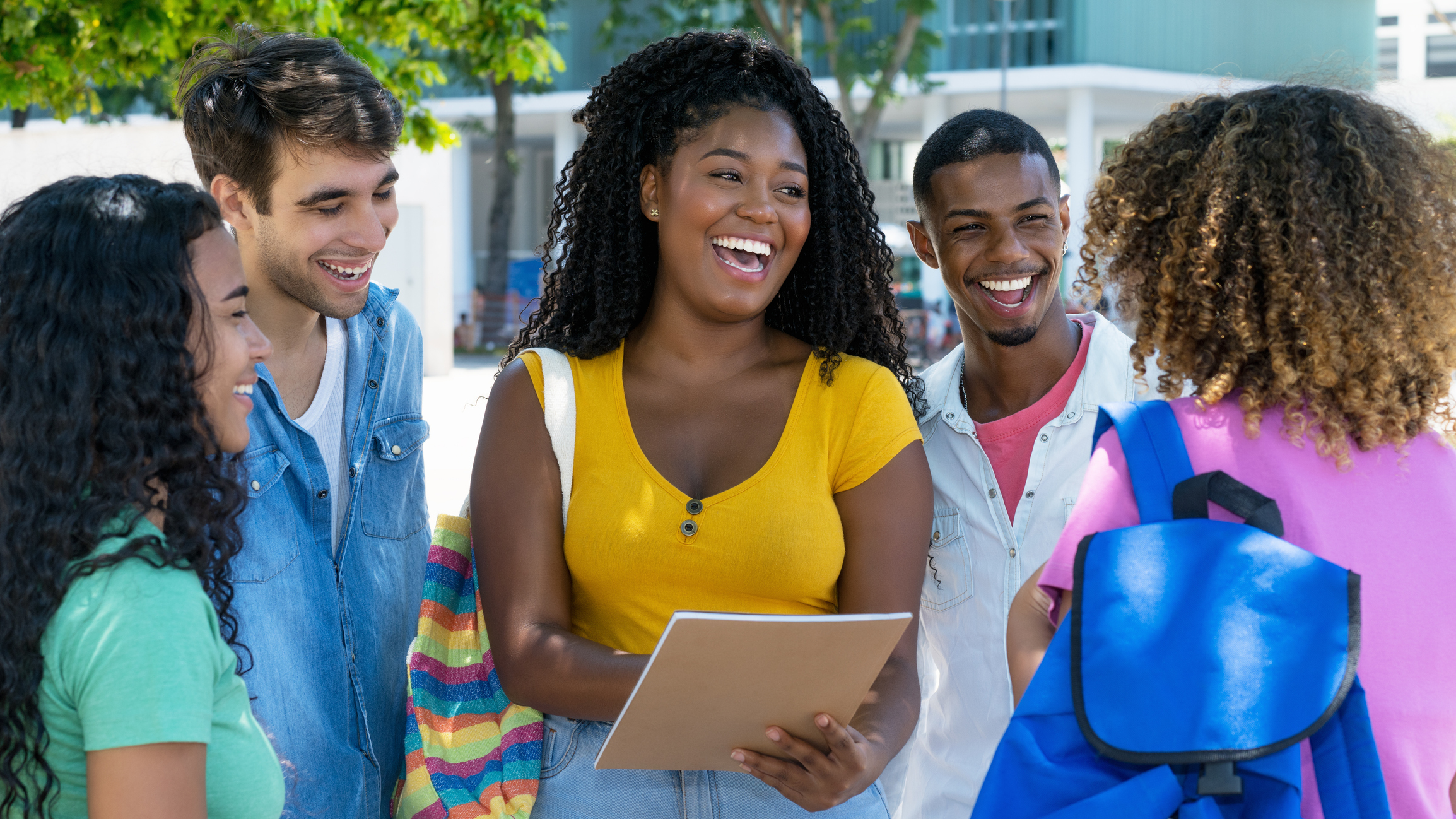 A group of five young adults stand outdoors, talking and smiling together. One woman holds a notebook while others listen attentively. Trees and buildings are visible in the background.