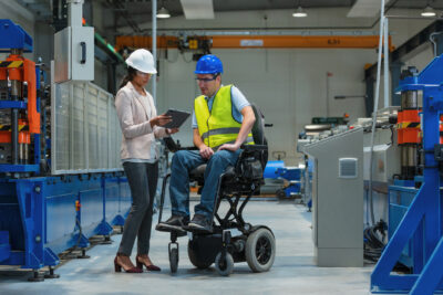 A woman in a white hard hat shows a tablet to a man in a blue hard hat and safety vest, who is sitting in a motorized wheelchair, inside a factory.
