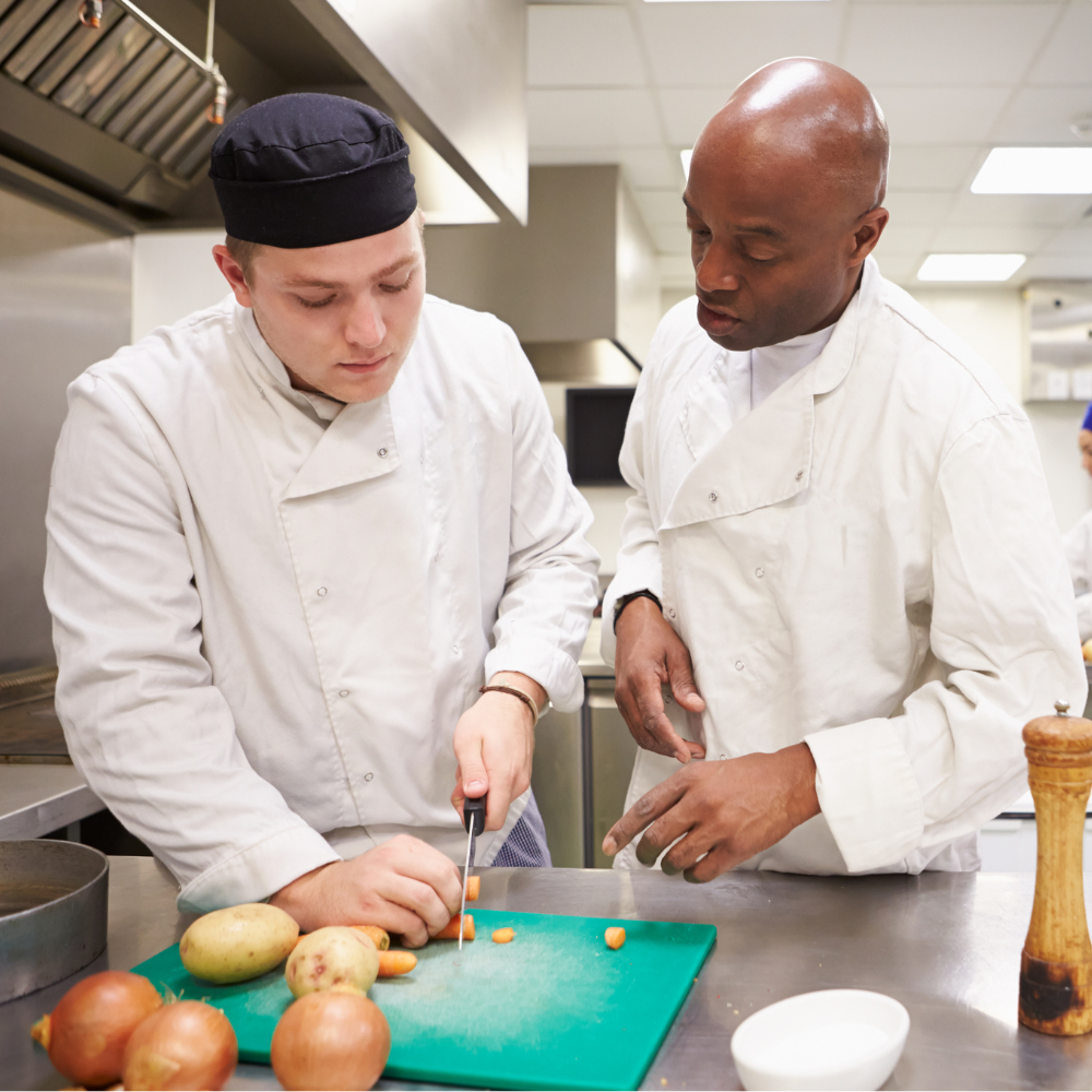 Two chefs in a commercial kitchen prepare food; one is slicing vegetables on a green board while the other observes and offers instruction.