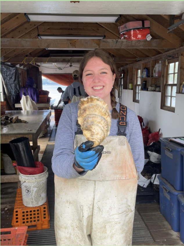 A person in work overalls and gloves stands indoors, smiling and holding a large oyster toward the camera inside a rustic workspace.