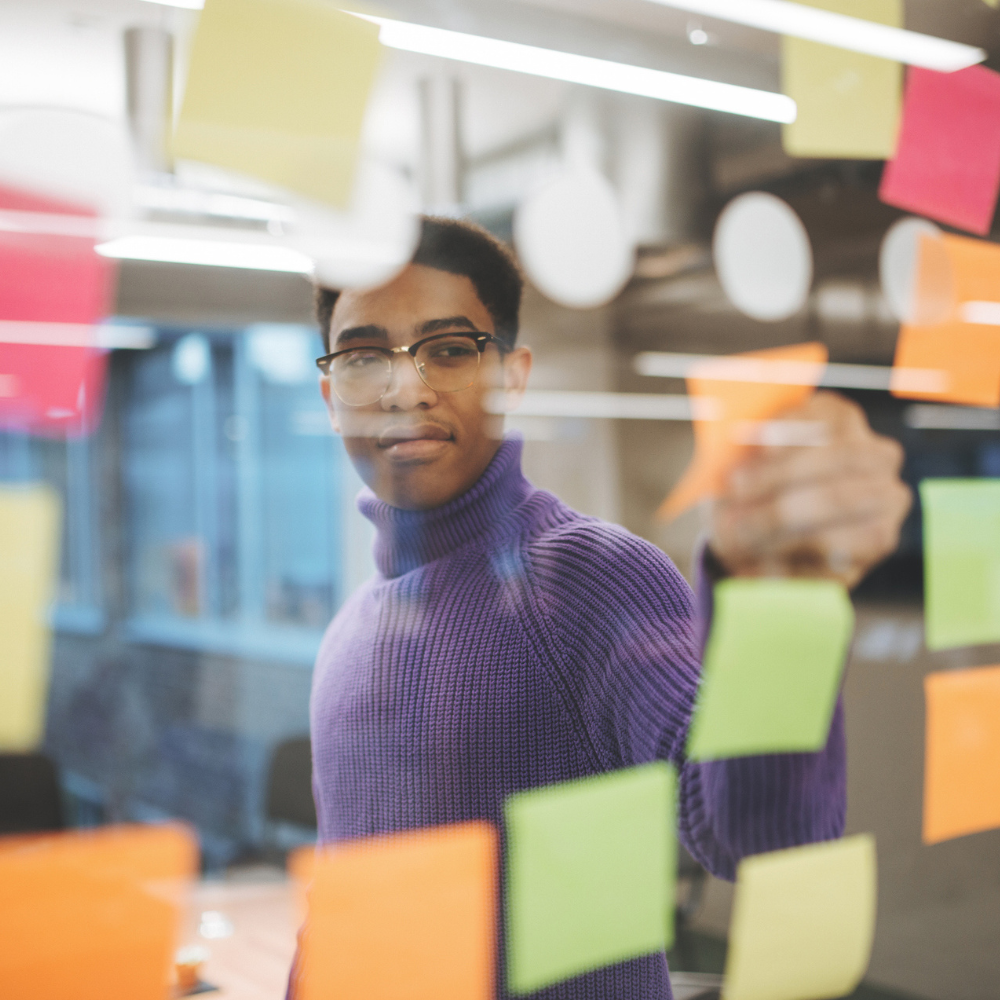 Person wearing glasses and a purple sweater places a sticky note on a glass wall covered with colorful sticky notes in a modern office setting.
