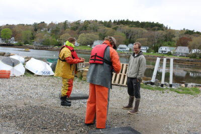 Two people in waterproof gear stand near a man in boots by the water, with boats, houses, and trees visible in the background.