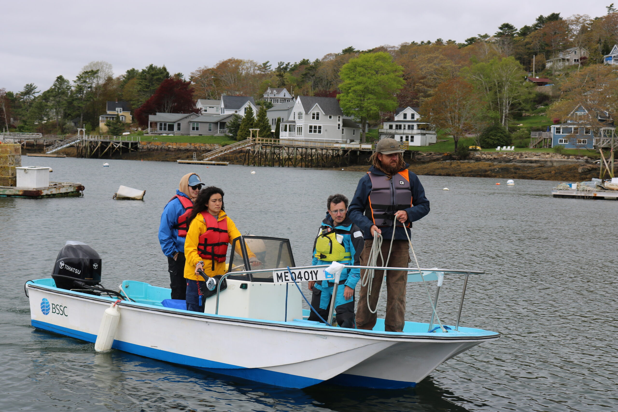 Four people wearing life jackets are on a small motorboat near the shore, with houses and trees visible in the background. One person stands at the bow holding a rope.