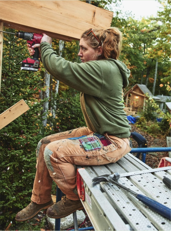 A person in work clothes sits on a metal scaffold outdoors using a power drill on a wooden beam, surrounded by trees and construction tools.